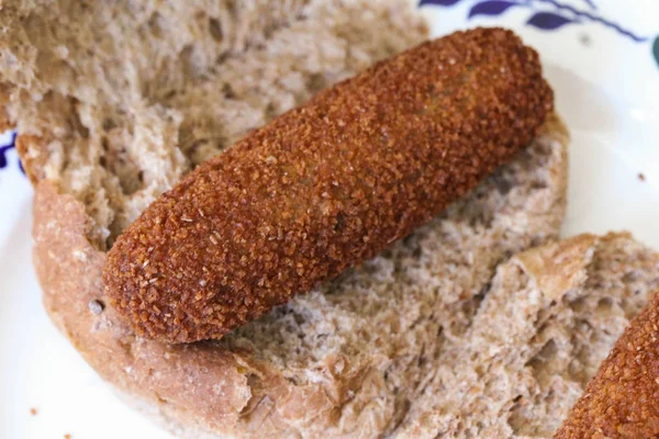 Crocchetta olandese fritta ripiena di manzo, chiamata "kroket", su un panino marrone, per pranzo - su piatto bianco su sfondo di legno — Foto Stock