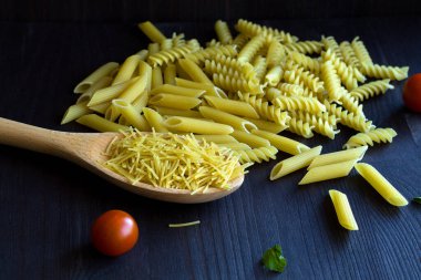 Fusilli çiğ makarna, siyah arka planda kiraz domatesli makarna. Masasında tahta kaşık ve fotokopi alanı olan farklı İtalyan makarnaları..