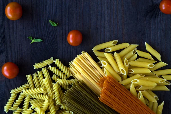 different types of Italian pasta on a wooden table with copy space. raw Spinach spashetti, wheat pasta and tomato spaghetti with fusilli, penne with cherry tomatoes on a black background. top view.