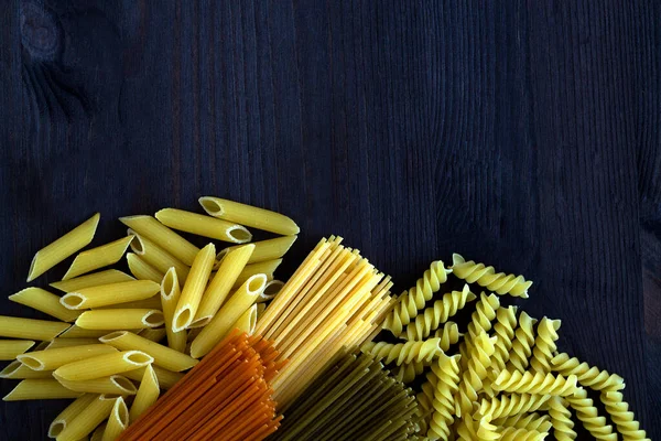 Italian Cuisine. different types of pasta on a wooden table with copy space. uncooked Spinach spashetti, wheat pasta and tomato spaghetti with  fusilli, penne on a black background. top view.
