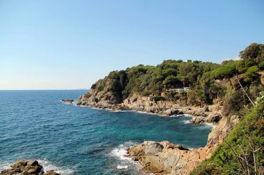 Denizin İspanya kıyıları. Yeşil çam ağaçları ve gökyüzünün altında dalgalar olan mavi denizli kayaların güzel manzarası. Akdeniz.