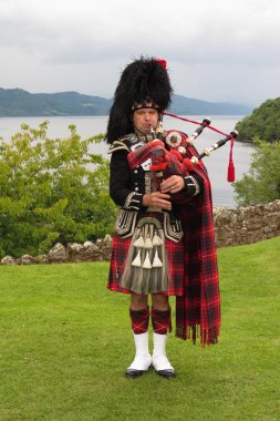 URQUHART CASTLE, SCOTLAND, UK - CIRCA AUGUST 2015: Scottish bagpiper dressed in traditional red and black tartan dress clipart