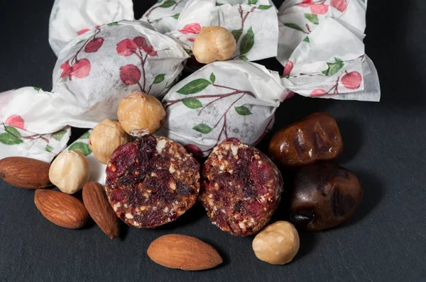 one cut candy  and wrapped handmade candies in colored paper are lying near the juicy dates, hazelnuts and almond nuts on a black background.