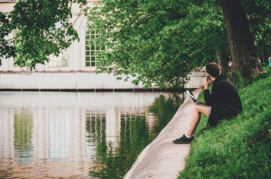 Yeşil çimenlerin üzerinde havuz dizlerinin üzerinde bir dizüstü bilgisayar ile oturup telefon olan genç adam. Yan görüntüle, kopya alanı.