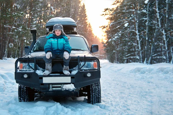 Έννοια του ταξιδιού χειμώνα. Χαριτωμένο αγόρι με μπλε χρώμα κάτω σακάκι κάθεται σε ένα προφυλακτήρα δύναμη ενός αυτοκινήτου στο δάσος του χειμώνα κατά τα χιονισμένα δέντρα. — Φωτογραφία Αρχείου