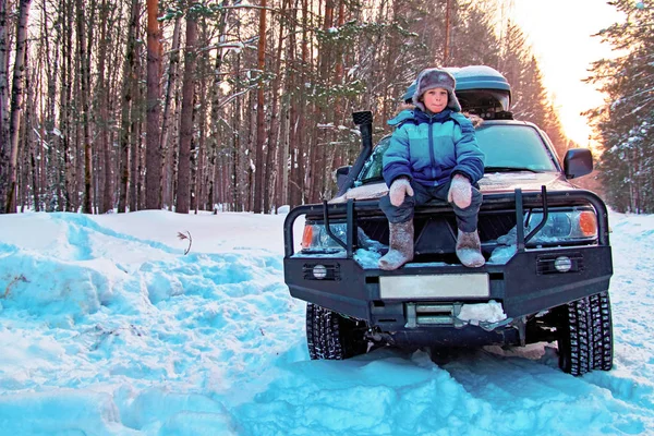 Concept of winter travel, recreation. Cute Boy in blue down jacket sits on a power bumper of a car in winter forest. Copy space.