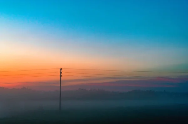 Avond de dikke mist over veld en bos. Landschap van de avondlucht met oranje en blauwe lucht en mist. Telegraaf pole in de mist. — Stockfoto