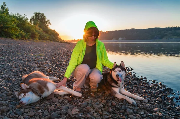 Chica en gafas de sol y cortavientos amarillos con perros husky en la orilla del río en el sol de la noche brillante. Retrato amante con perros . —  Fotos de Stock