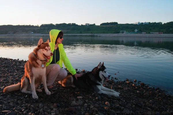 Ragazza in occhiali da sole e giacca a vento gialla con cani husky sulla riva del fiume. Concetto proprietario a piedi con animali domestici nel parco . — Foto Stock