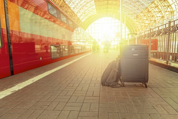 Mochila y maleta con ruedas en la estación de tren a la luz del sol. Equipaje a la espera del tren. Puesta de sol, concepto de viaje . — Foto de Stock