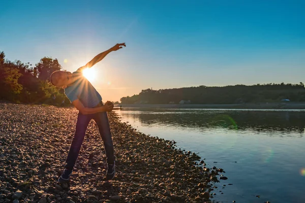 Boy (11-12 years) throws stones at the river on the background summer sunset. Sun illuminates the silhouette of a child in blue t-shirt and jeans.