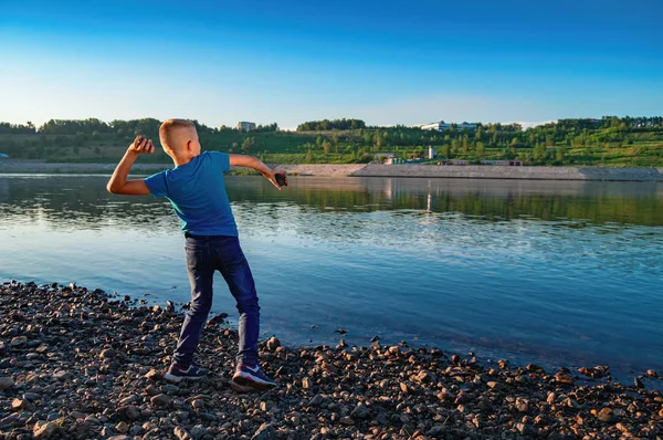 Boy (11-12 years) throws stones at the river on the background summer sunset. Child in blue t-shirt and jeans. Back view.