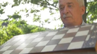 Yaşlı, beyaz saçlı adamın ön görüntüsü bahçedeki satranç oyununu açıyor ve siyah figürleri yan yana koyuyor..