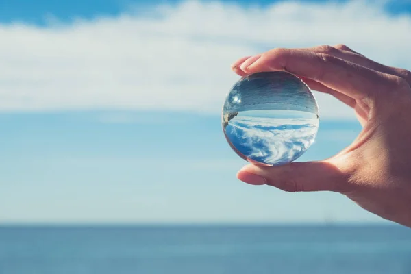 Woman's hand holding a crystal ball, looking through to the ocean and sky. Creative photography, crystal ball refraction — Stock Photo, Image