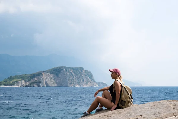 Una joven con el pelo rubio se sienta en la cima de una montaña y mira el mar y la playa. Relájate y viaja por la naturaleza . — Foto de Stock