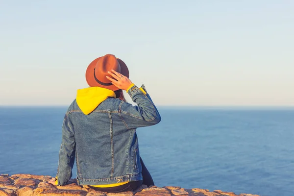 Nahaufnahme Rückenansicht einer Frau in Jeansjacke und Hut, die sitzt und auf blauen Ozean und Himmel blickt. — Stockfoto