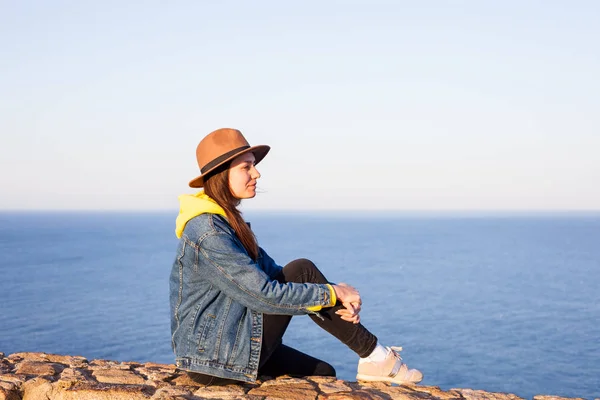 Vista posterior de cerca de la mujer en chaqueta vaqueros y sombrero sentado y mirando el océano azul y el cielo . — Foto de Stock