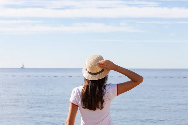 Hermosa chica mira al mar. Chica joven en un sombrero mirando a un mar tranquilo y cielos azules vista trasera . — Foto de Stock