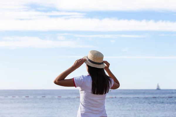 Hermosa chica mira al mar. Chica joven en un sombrero mirando a un mar tranquilo y cielos azules vista trasera . — Foto de Stock