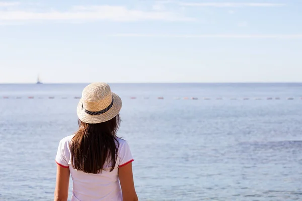 Hermosa chica mira al mar. Chica joven en un sombrero mirando a un mar tranquilo y cielos azules vista trasera . — Foto de Stock
