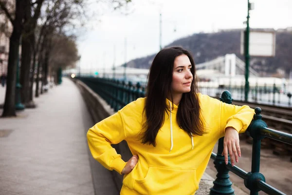 Jovem elegante em amarelo com capuz posando nas ruas da cidade. Mulher de estilo urbano — Fotografia de Stock