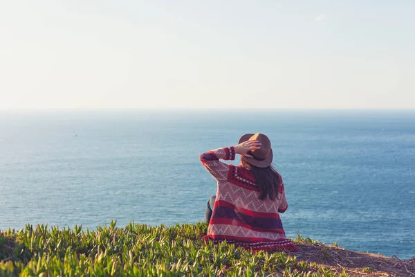 Primer plano vista trasera de la mujer en ropa de viaje y sombrero sentado y mirando el océano azul y el cielo . — Foto de Stock