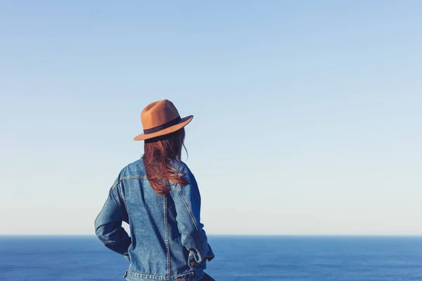 ジーンズジャケットと帽子をかぶった女性のクローズアップバックビュー立ち、青い海と空を眺めながら. — ストック写真