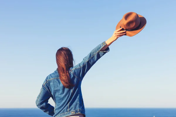 Vista trasera de cerca de la mujer en chaqueta vaquera sosteniendo el sombrero y de pie y mirando el océano azul y el cielo . — Foto de Stock