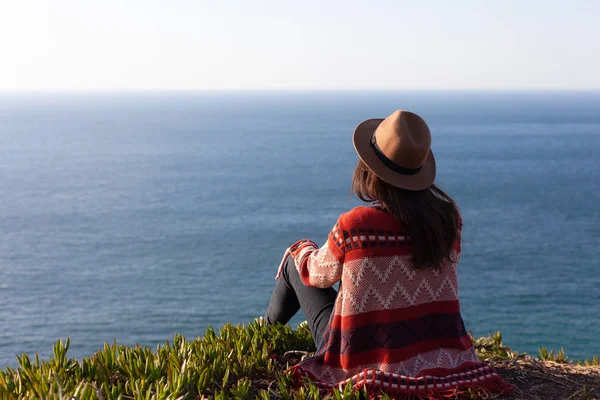 Primer plano vista trasera de la mujer en ropa de viaje y sombrero sentado y mirando el océano azul y el cielo . — Foto de Stock