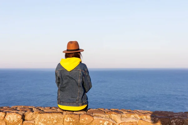 Nahaufnahme Rückansicht einer Frau in Reisekleidung und Hut, die sitzt und auf blauen Ozean und Himmel blickt. — Stockfoto