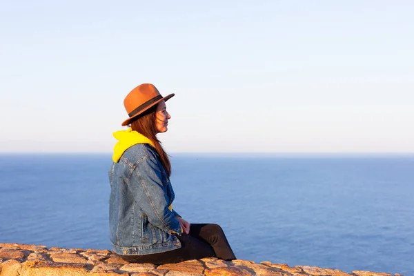 Frau in Reisekleidung und Hut sitzt und auf blauen Ozean und Himmel blickt. — Stockfoto