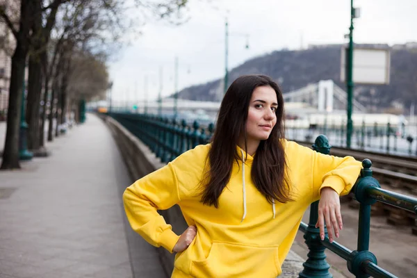 Jovem elegante em amarelo com capuz posando nas ruas da cidade. Mulher de estilo urbano — Fotografia de Stock