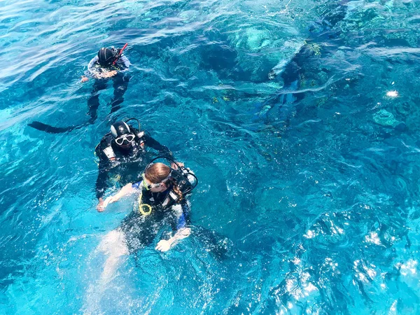 Three underwater divers in black scuba diving suits, a man and a woman with oxygen bottles sink under the transparent blue water in the sea, the ocean in a tropical paradise warm resort.