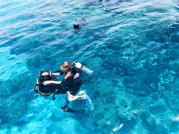 Three underwater divers in black scuba diving suits, a man and a woman with oxygen bottles sink under the transparent blue water in the sea, the ocean in a tropical paradise warm resort.