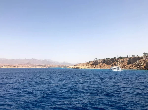 Uzak güzel tropikal kahverengi taş dağlar ve çeşitli binalar, sahildeki gemi ve mavi tuz Azure deniz, okyanus mavi gökyüzüne karşı deniz manzarası.