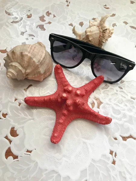 A beautiful red starfish and black sunglasses lie decorated with seashells on a white cloth background. Marine tropical summer background
