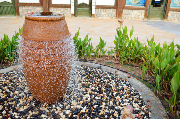 A beautiful little fountain in the form of a brown vase, a jug with falling drops of water on colored stones standing in a flower bed with green plants.