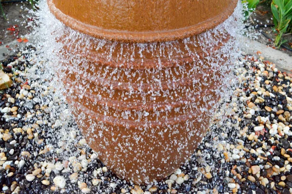 Una bella fontana a forma di vaso marrone, una brocca con gocce d'acqua cadenti su pietre colorate in piedi in un'aiuola con piante verdi — Foto Stock