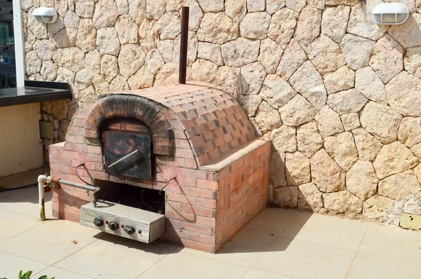 Un gran horno de parrilla industrial de piedra marrón profesional con un tubo para cocinar alimentos de una baldosa de piedra sobre un fondo de pared de piedra en la calle al aire libre — Foto de Stock
