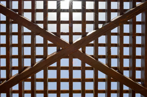 Textura de una celosía abstracta de madera marrón con celdas cuadradas con agujeros de tableros de vigas de madera dispuestas verticalmente horizontalmente diagonalmente sobre el fondo del sol y el cielo. El trasfondo — Foto de Stock
