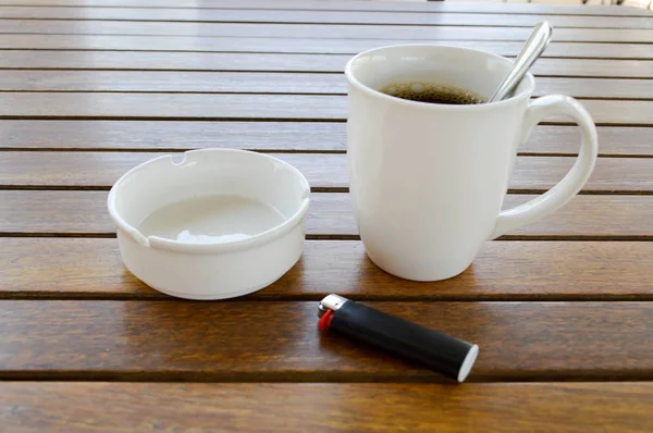 Uma xícara de cerâmica branca com uma manhã refrescante café quente com chá e chá colher de chá brilhante e um cinzeiro vazio com um isqueiro preto fica em uma mesa de madeira em um café — Fotografia de Stock