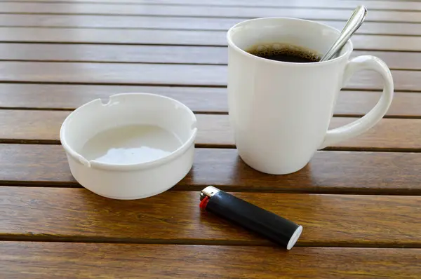 Uma xícara de cerâmica branca com uma manhã refrescante café quente com chá e chá colher de chá brilhante e um cinzeiro vazio com um isqueiro preto fica em uma mesa de madeira em um café — Fotografia de Stock