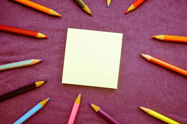 Разноцветные, яркие, разноцветные карандаши расположены вокруг листа бумаги слева и место для текста на фоне фиолетовой ткани — стоковое фото
