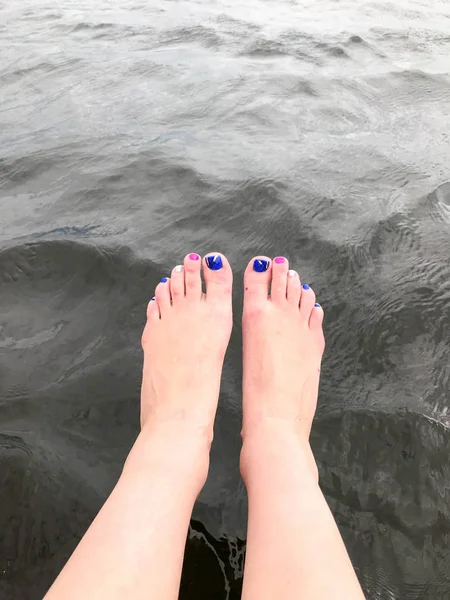 Belos pés femininos dos pés com os dedos com uma manicure de moda multi-colorida são deixados pendurados na água preta escura fria — Fotografia de Stock