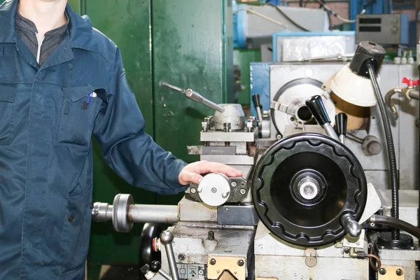 Um homem trabalhando em um manto, macacão fica ao lado de um torno industrial para cortar, transformando facas de metais, madeira e outros materiais, girando, fazendo detalhes e peças de reposição na fábrica — Fotografia de Stock