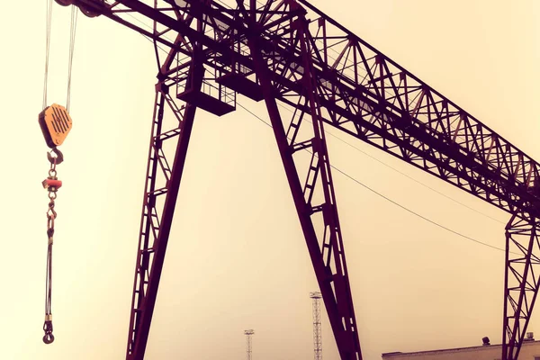 Grúa pórtico metálica grande de hierro industrial con un gancho montado en los soportes para levantar y transportar cargas pesadas, moviéndose a lo largo de los rieles de la fábrica. La grúa es de tipo puente —  Fotos de Stock