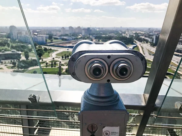 Viewing coin stationary binoculars against a white-blue sky in profile. Gray metal binoculars on the viewing platform at a height. From the roof overlooking the beautiful green city