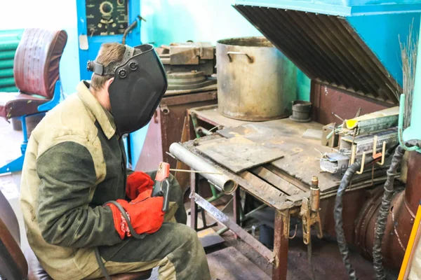 Um trabalhador masculino um soldador em uma máscara protetora solda um tubo de metal em uma estação de soldagem em uma oficina em uma planta metalúrgica em uma instalação de reparo — Fotografia de Stock