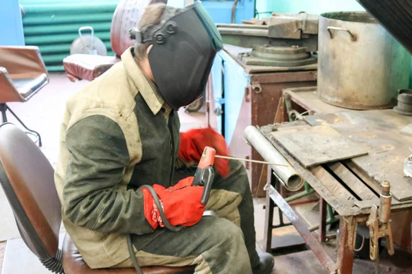 Um trabalhador masculino um soldador em uma máscara protetora solda um tubo de metal em uma estação de soldagem em uma oficina em uma planta metalúrgica em uma instalação de reparo — Fotografia de Stock