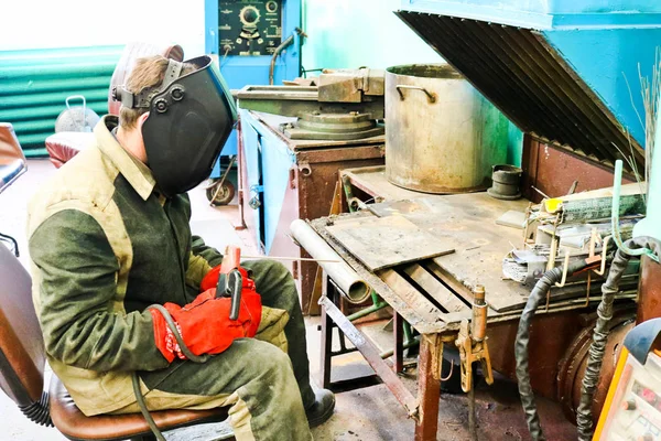 Um trabalhador masculino um soldador em uma máscara protetora solda um tubo de metal em uma estação de soldagem em uma oficina em uma planta metalúrgica em uma instalação de reparo — Fotografia de Stock